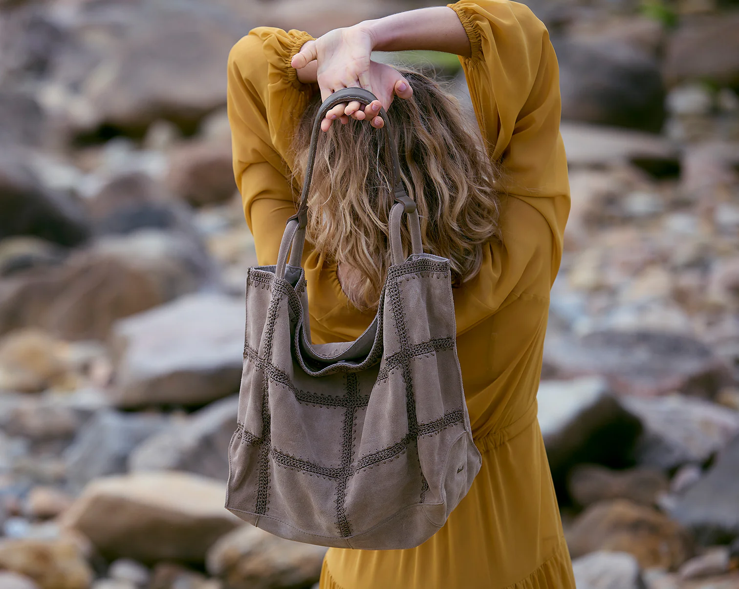 In a picture from The Sak, a woman in a yellow sweater holds a crocheted bag behind her back.